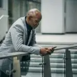Confident businessman checks his mobile phone while leaning on railing in cityscape.