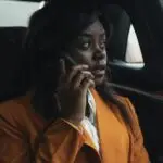 A businesswoman in an orange suit talking on the phone inside a car, portraying leadership and professionalism.
